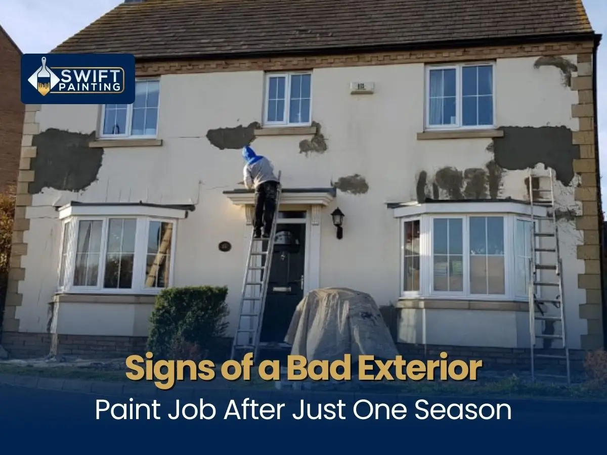 A two-story house with patches of peeling and damaged exterior paint being repaired by a worker on a ladder.