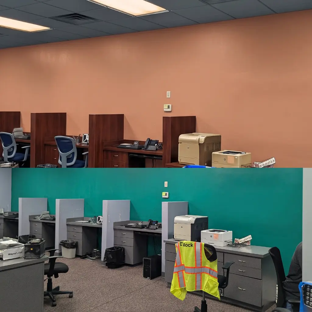 Open plan corporate office space featuring multiple grey cubicle workstations against a teal green wall, with office chairs, printers, and a yellow safety vest hanging on a chair.